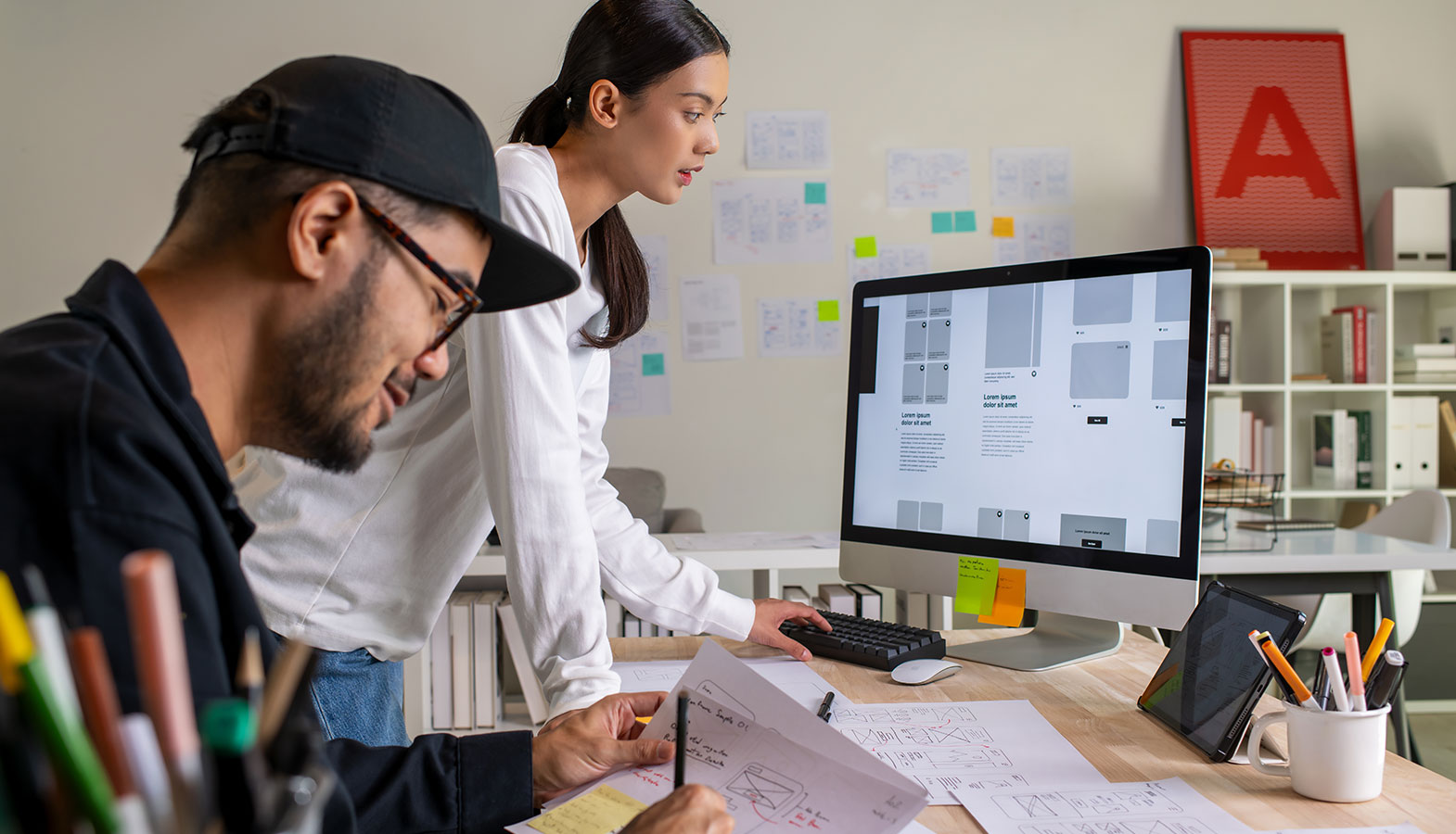 A designer is intently reviewing website wireframes displayed on a large monitor, with various text blocks and image elements visible on the screen. The scene captures the process of optimizing the largest contentful paint (LCP) to improve the web page's performance metrics.